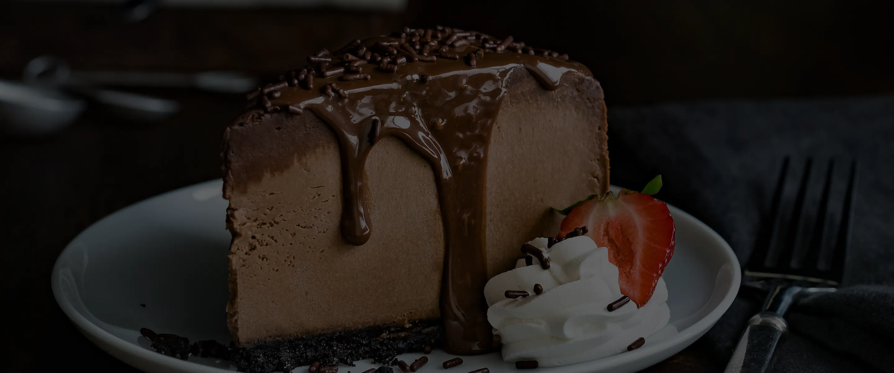 A slice of chocolate cheesecake with chocolate drizzle, whipped cream, sprinkles, and a strawberry on a white plate with a fork beside it.